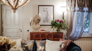 Una habitación con un sofá azul, una escultura de busto, un dibujo enmarcado, un jarrón con flores y una ventana con cortinas.