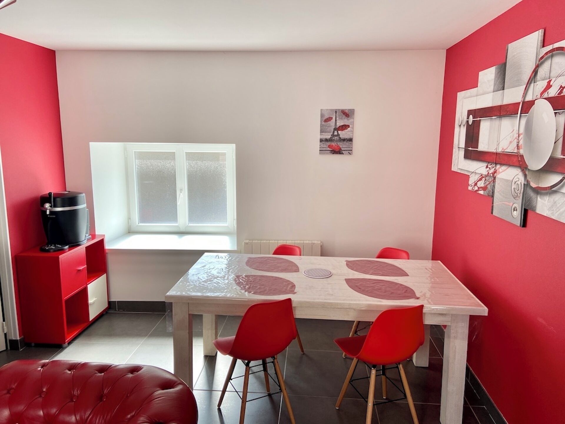 A dining area with a white table, red chairs, a red leather bench, a window, and a red wall with a picture.