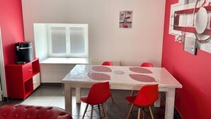 Un coin repas avec une table blanche, des chaises rouges, un banc en cuir rouge, une fenêtre et un mur rouge orné d’un tableau.