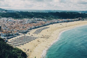 Un paysage urbain côtier avec une plage bordée de rangées de chaises longues et de parasols, des bâtiments aux toits recouverts de tuiles rouges et une colline en arrière-plan.