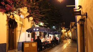 Une ruelle étroite pavée, éclairée la nuit, avec des tables et des chaises installées à l’extérieur sous des parasols, une enseigne indiquant « IRGUESINHA » et un banc en bois avec une plaque.