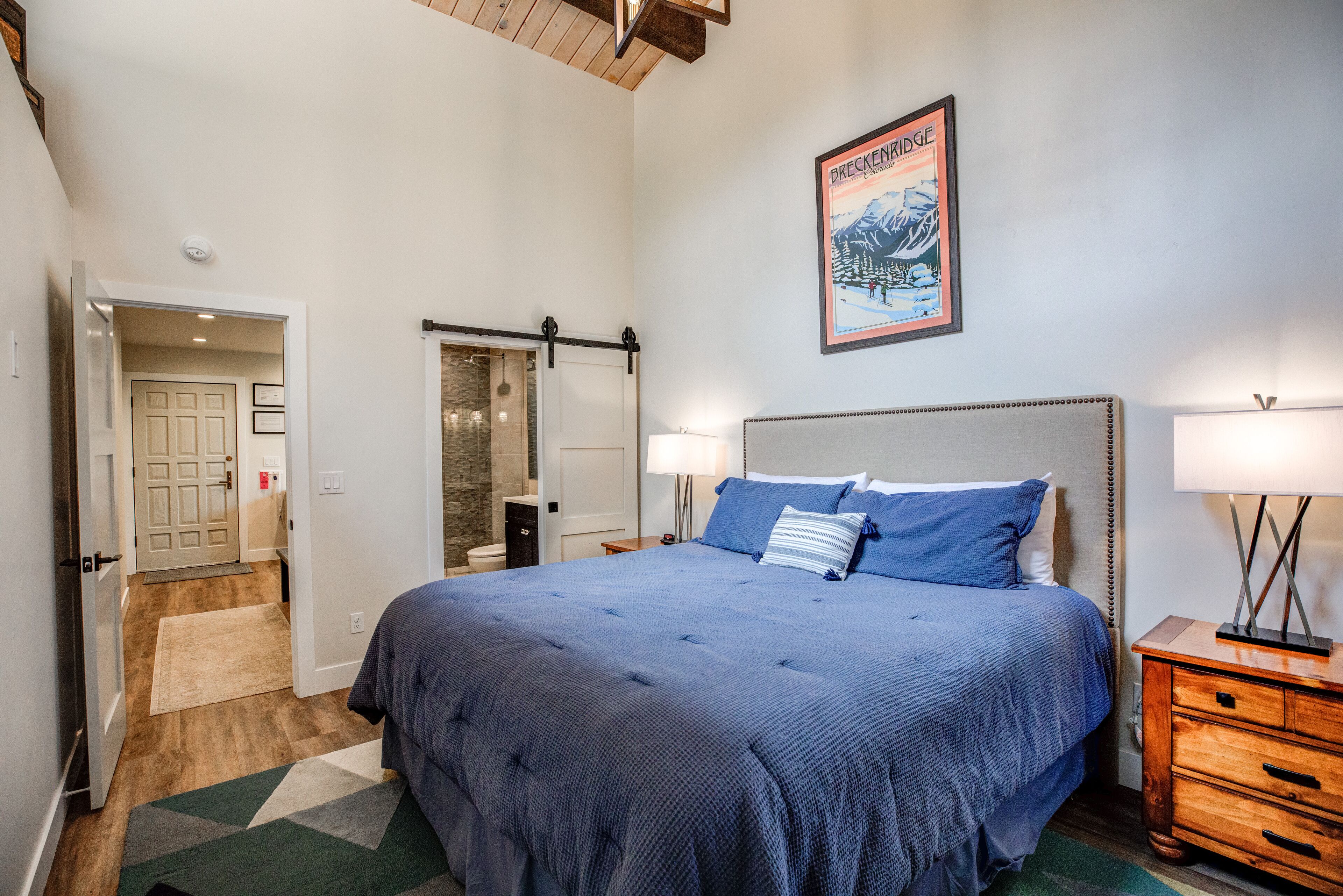 Een slaapkamer met een groot bed, een houten nachtkastje, een lijst met een afbeelding aan de muur en een deur naar een andere kamer.