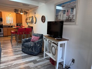 A living room with a television, a patterned armchair, a red cushion, and a kitchen in the background.