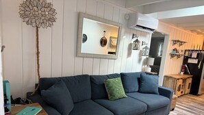 Una sala moderna con un sofá azul oscuro, un cojín estampado en verde, una mesita de madera con un cuaderno y una pieza decorativa en la pared.