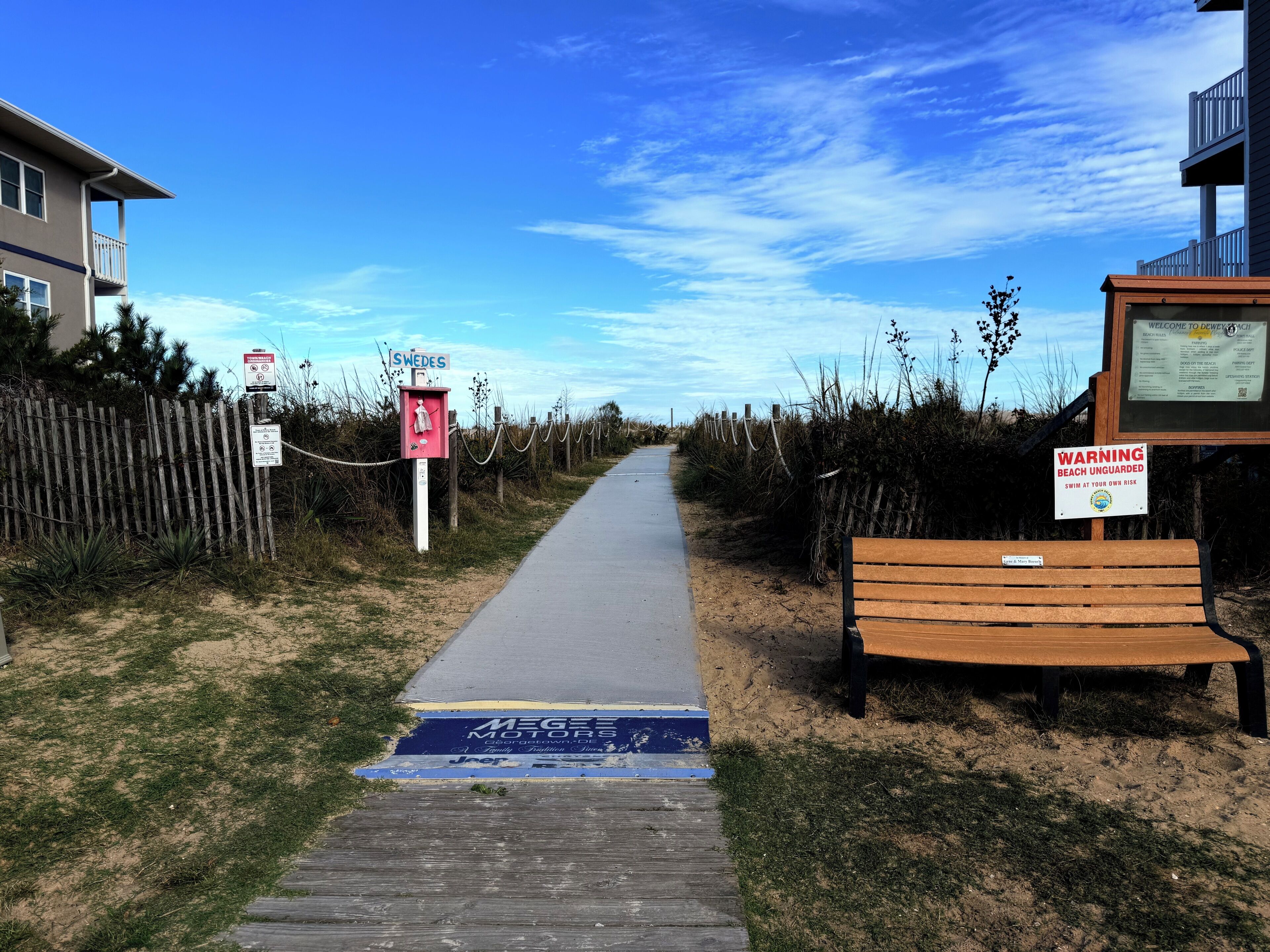 Un sendero que lleva a una playa con un banco de madera y señales de advertencia.
