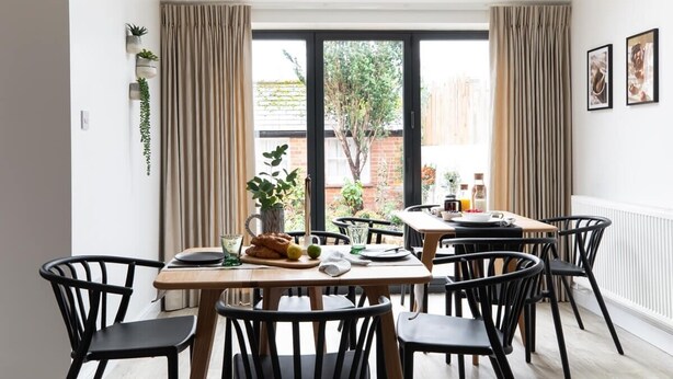 A dining area with a table set for four, surrounded by black chairs, a window with curtains, and a view of a brick wall and greenery outside. - The Oxford Lodge | Oxford Holiday Let | 6 Beds & Sleeps 12 (Oxford)