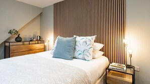 A modern bedroom with a sloped ceiling, a wooden headboard, and a bedside table with a book and a lamp. - The Oxford Lodge | Oxford Holiday Let | 6 Beds & Sleeps 12 (Oxford)