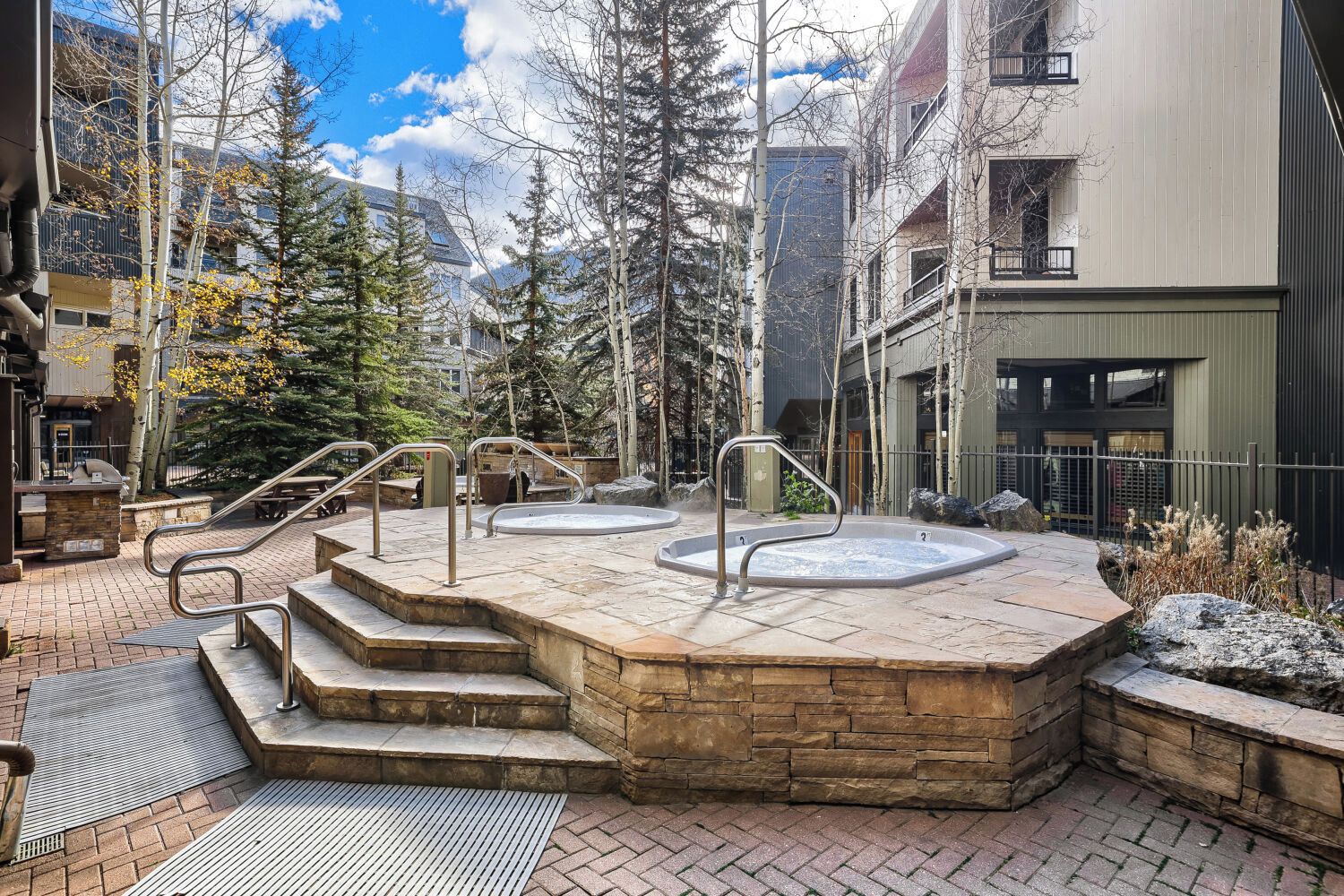 A spa area with stone steps leading to hot tubs, surrounded by trees and modern buildings.