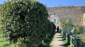 Un sentiero sterrato conduce a un edificio in pietra con una recinzione verde e un siepe sul lato sinistro.