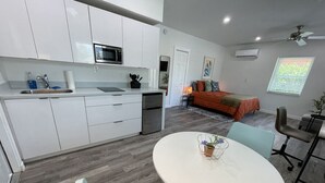 Eine moderne Küche mit weißen Schränken, einer Mikrowelle und einem Spülbecken. Es gibt eine Essecke mit einem runden Tisch und Stühlen. Im Hintergrund ist ein Bett mit einer orangefarbenen Decke zu sehen.