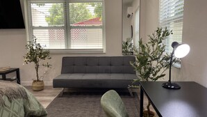 A modern bedroom with a grey sofa, a small desk with a lamp, two potted plants, and a large window with blinds. - 526 · Beautiful and Spacious studio - Casa Habana (Little Habana)
