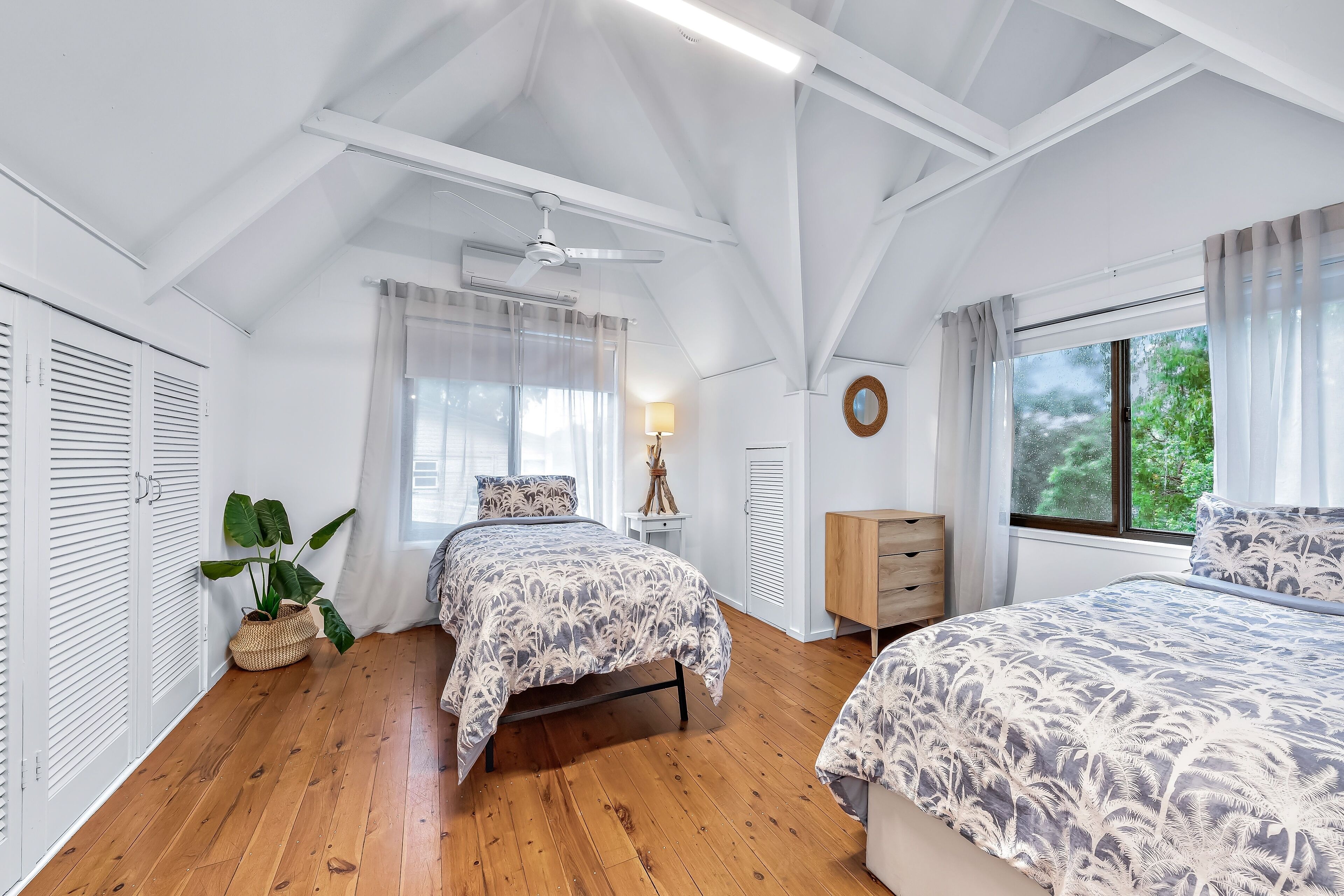 Une chambre avec deux lits, un plancher en bois, des murs blancs, un ventilateur de plafond et une fenêtre avec des rideaux.