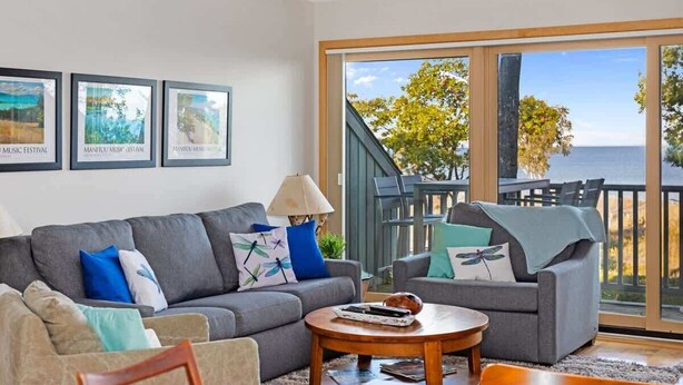 A living room with a grey sofa, a coffee table, and a view of the sea through large glass doors. - Dragonfly at the Homestead (Glen Arbor)