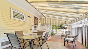 A covered patio with a glass table and metal chairs, a white fence, and a yellow house in the background.