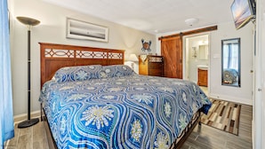 A bedroom with a large bed, a nightstand, a lamp, a TV, and a bathroom visible through an open door.
