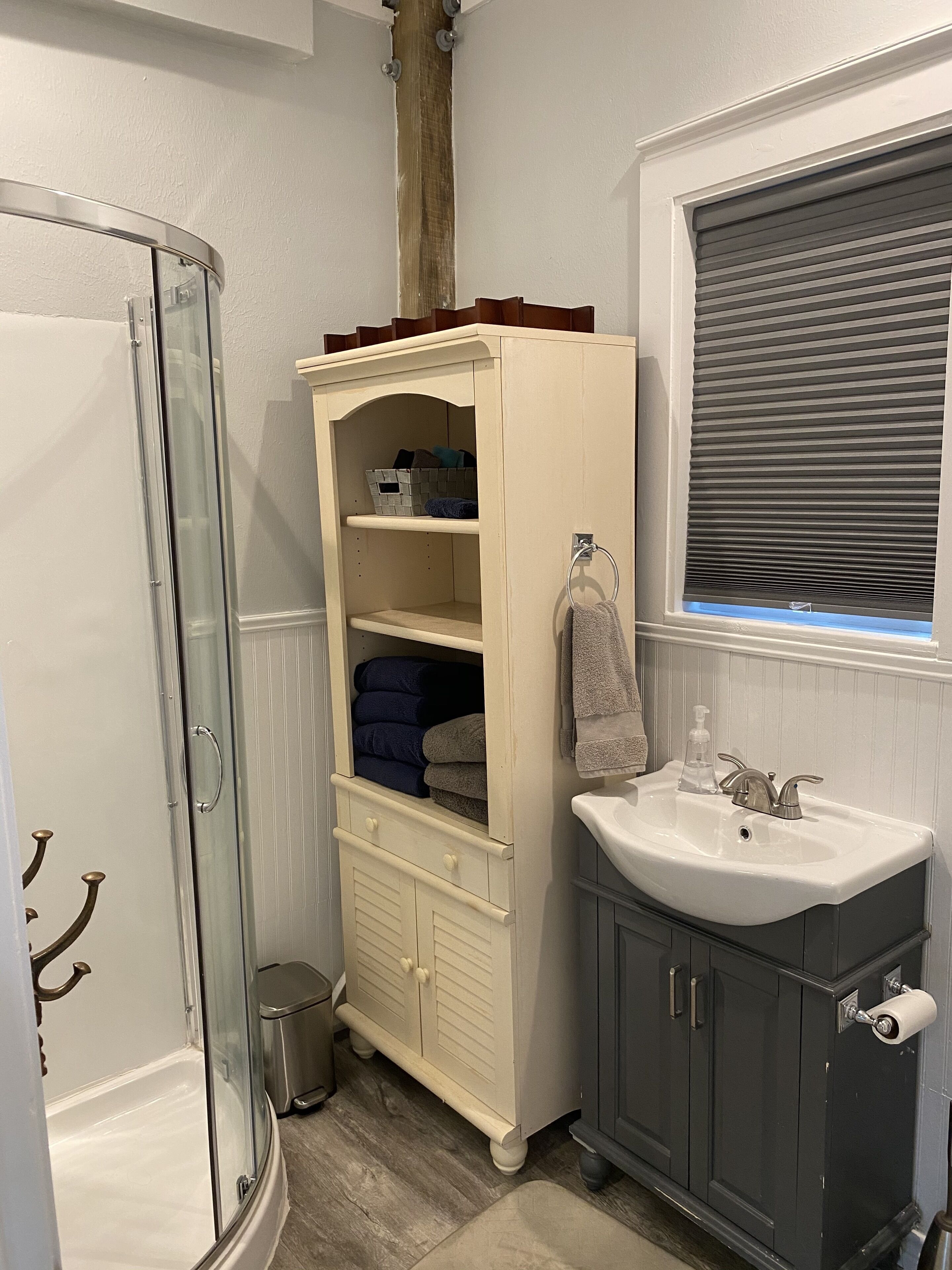 A bathroom with a glass shower enclosure, a beige vanity with a sink, and a window with blinds.