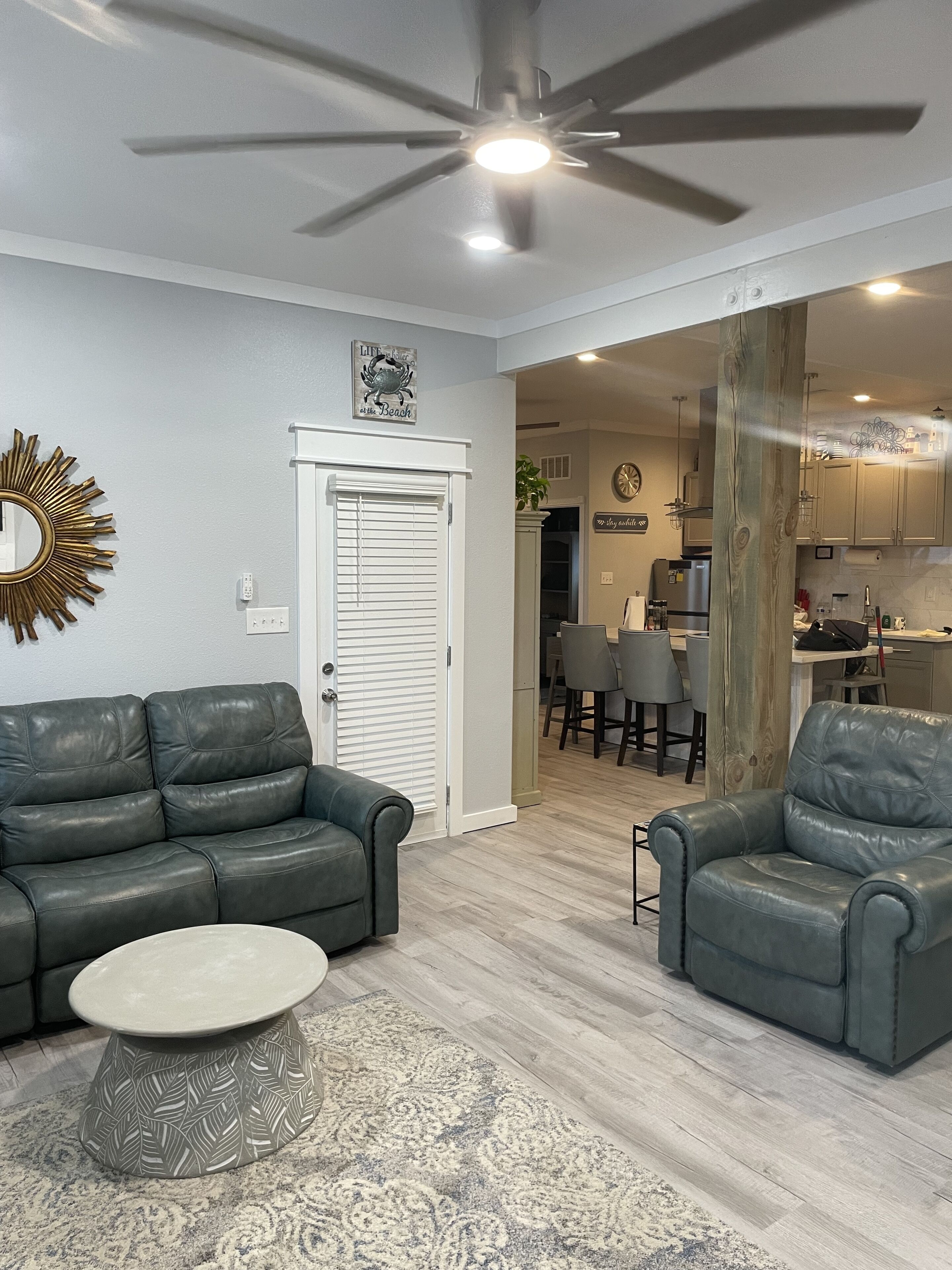Un salon avec deux canapés en cuir, une table basse et un tapis à motifs. On peut voir un ventilateur de plafond, et une porte donne accès à une cuisine.
