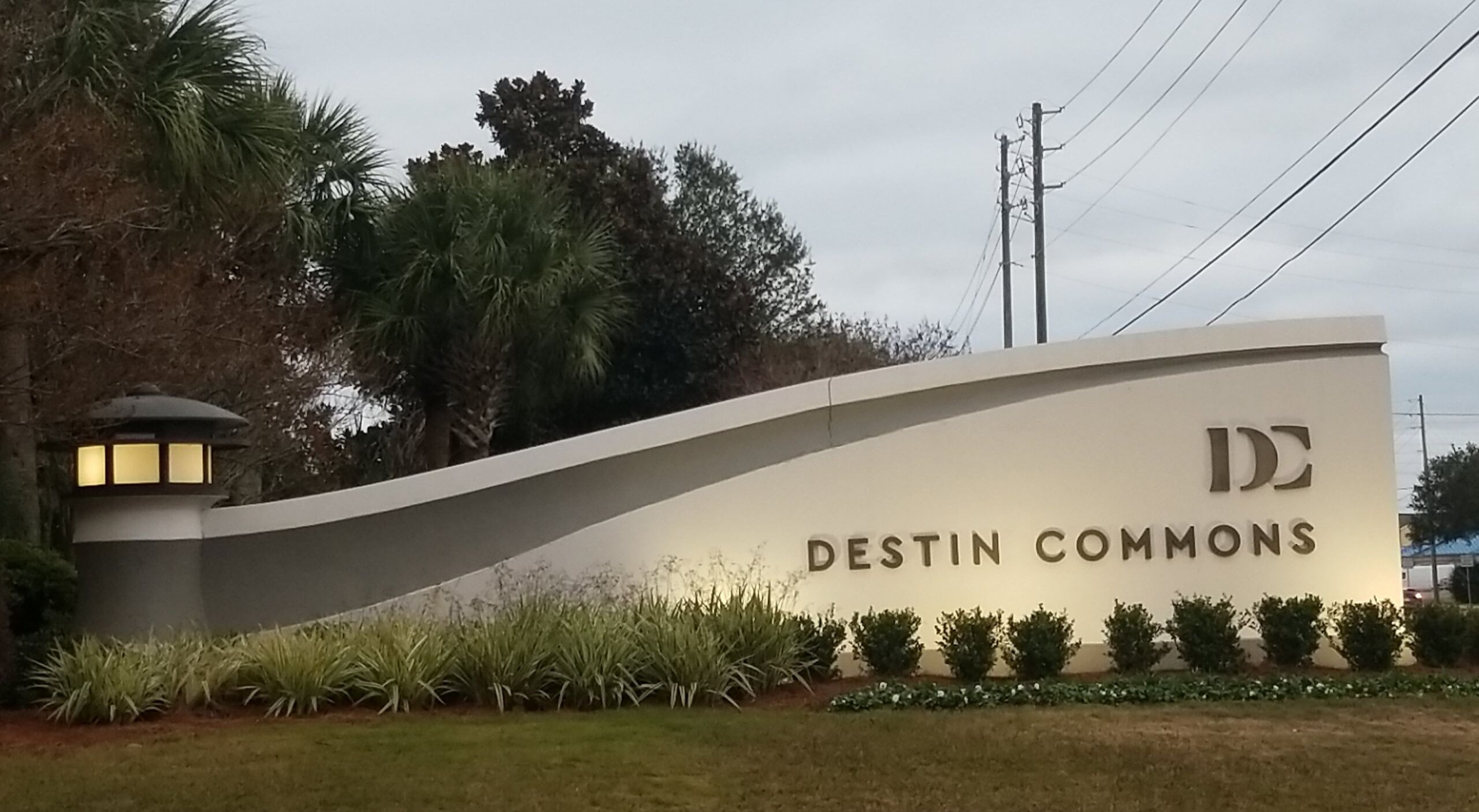 A sign for Destin Commons with a curved design, palm trees, and a lamp post.