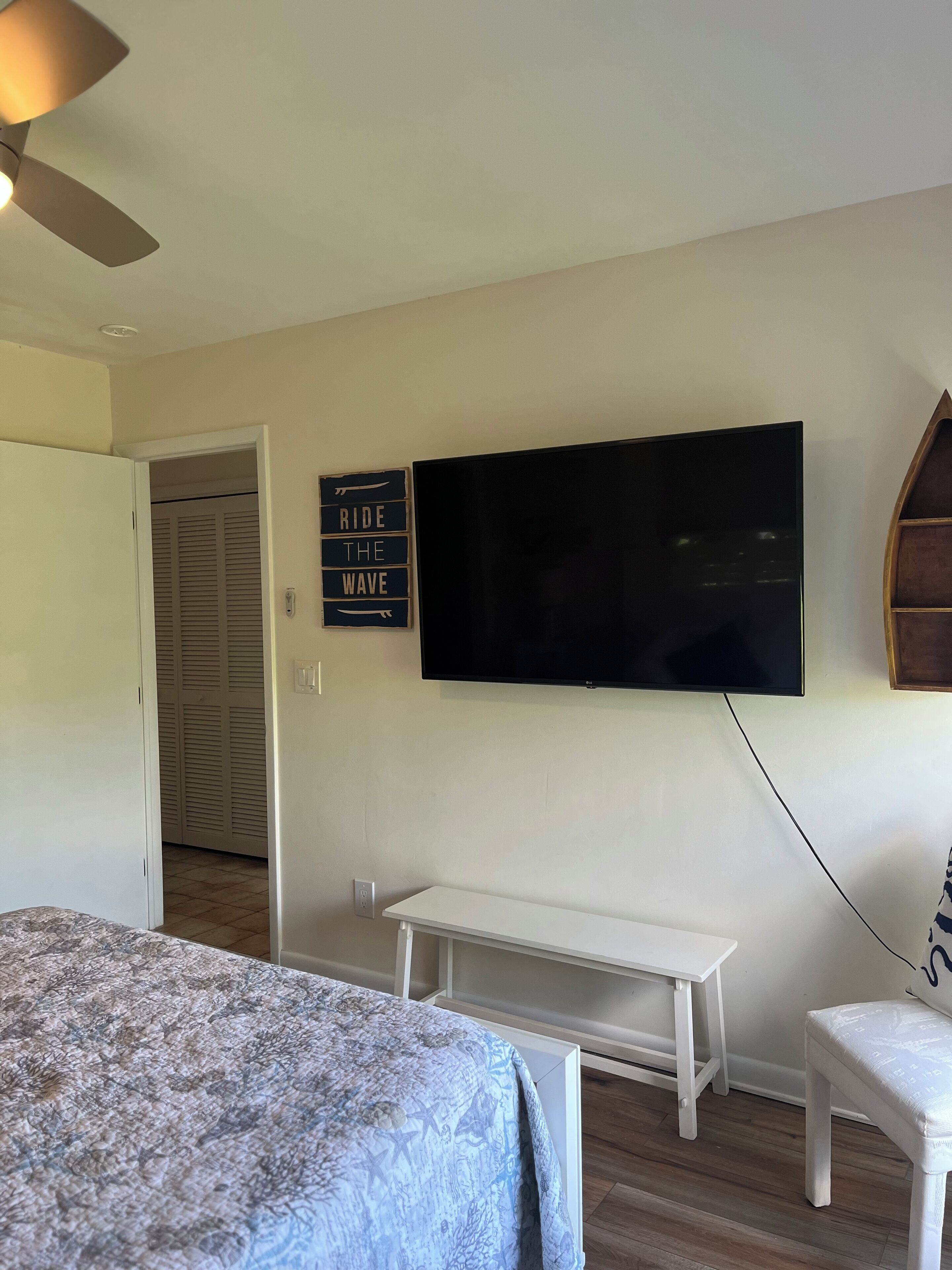 A bedroom with a bed, a TV mounted on the wall, a bench, and a sign that says "RIDE THE WAVE".
