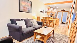 A living room with a sofa, a coffee table, and a patterned rug. There is a dining area with wooden chairs and a table. - Two Storey Cozy Chalet W/2BR&2BA Balcony Mountain Views/By Banff Gate / Sleep 6 (Harvie Heights)