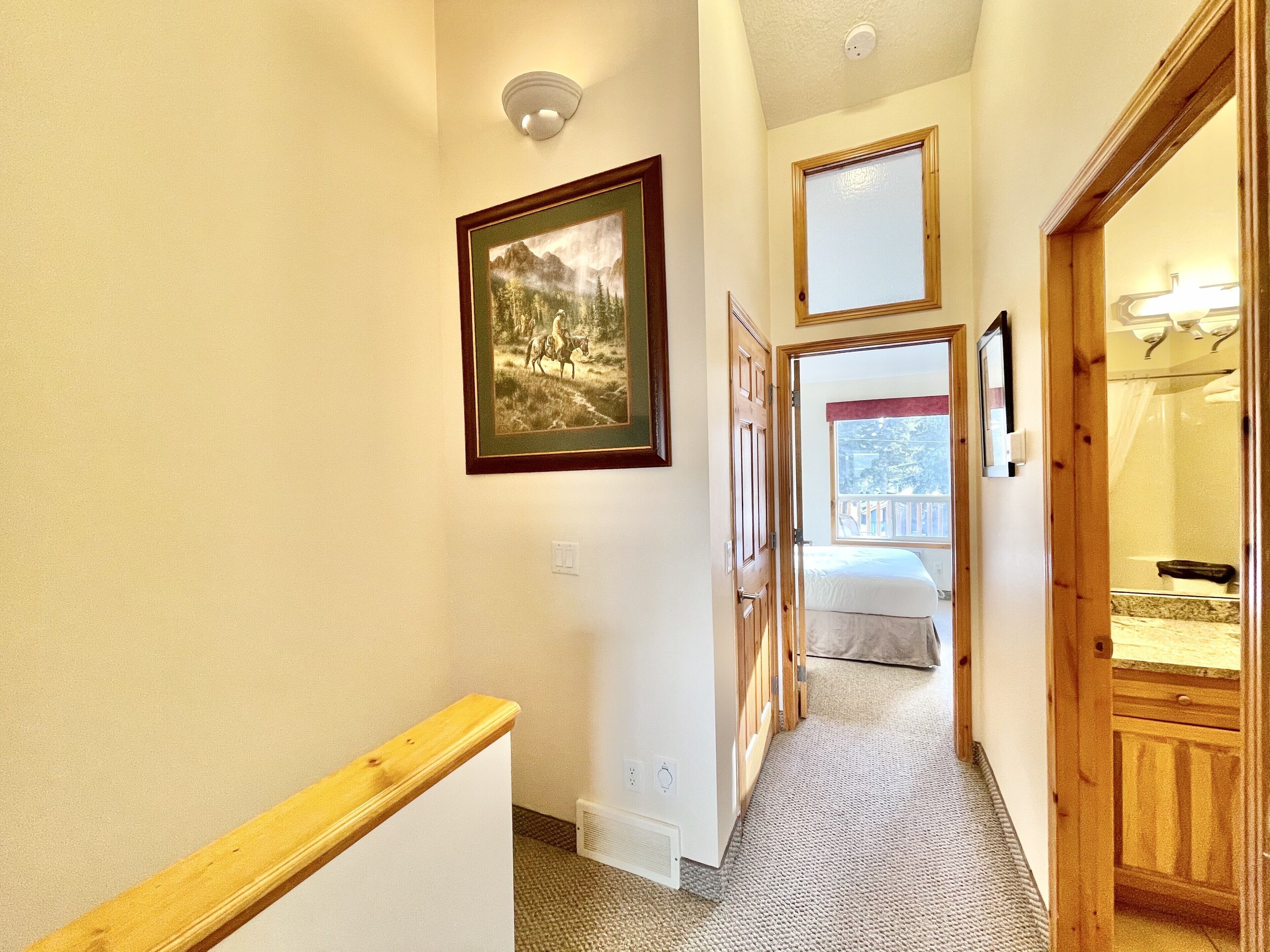 A hallway with a wooden handrail, a framed picture on the wall, and a bed visible through an open door.