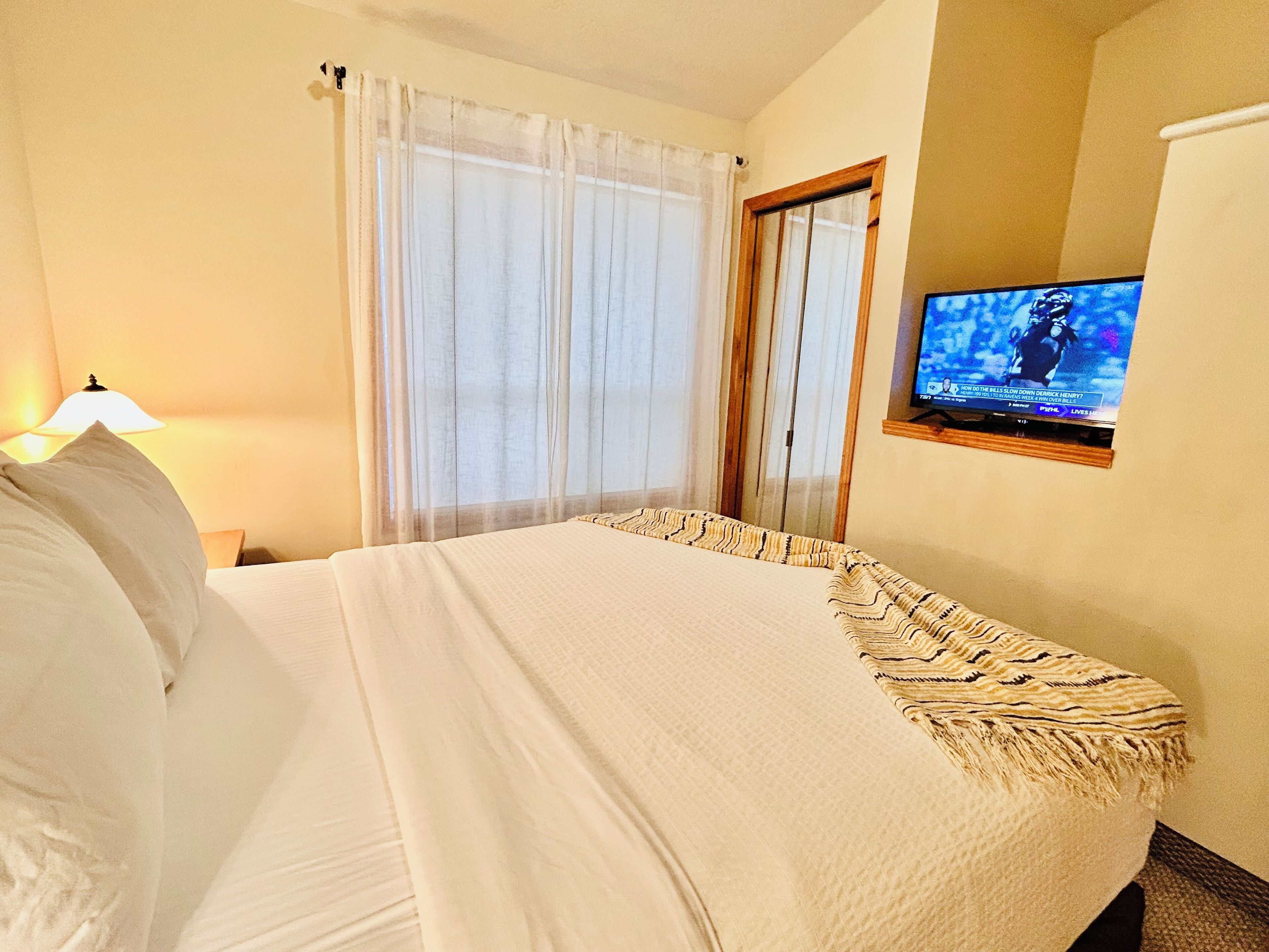 A hotel room with a bed, a TV mounted on the wall, a window with curtains, and a lamp on the side table.