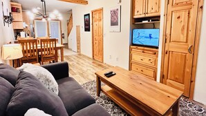 A living room with a sofa, wooden cabinets, a TV, and a dining area with chairs and a table. - Two Storey Cozy Chalet W/2BR&2BA Balcony Mountain Views/By Banff Gate / Sleep 6 (Harvie Heights)