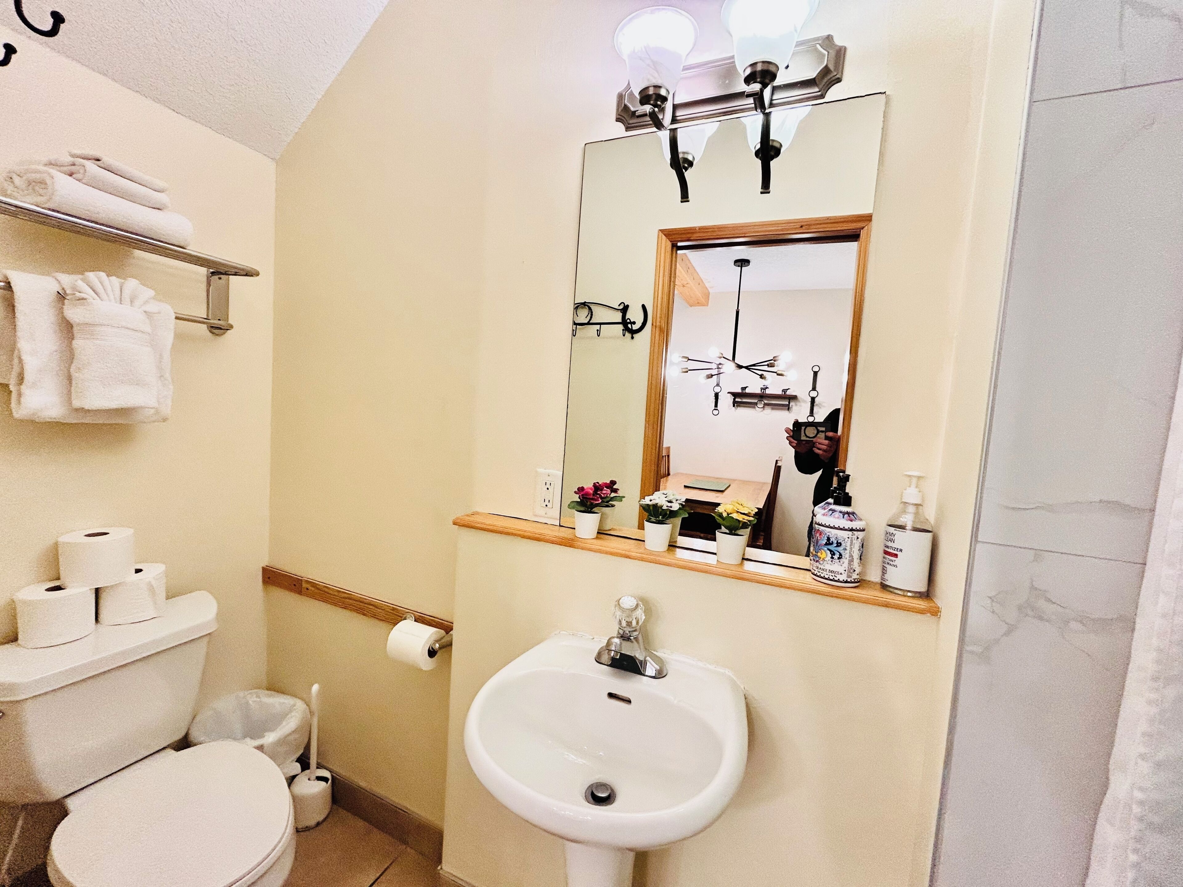 A bathroom with a toilet, sink, and mirror. There are towels on a rack, a trash can, and a shelf with toiletries.