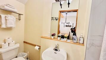 A bathroom with a toilet, sink, and mirror. There are towels on a rack, a trash can, and a shelf with toiletries.