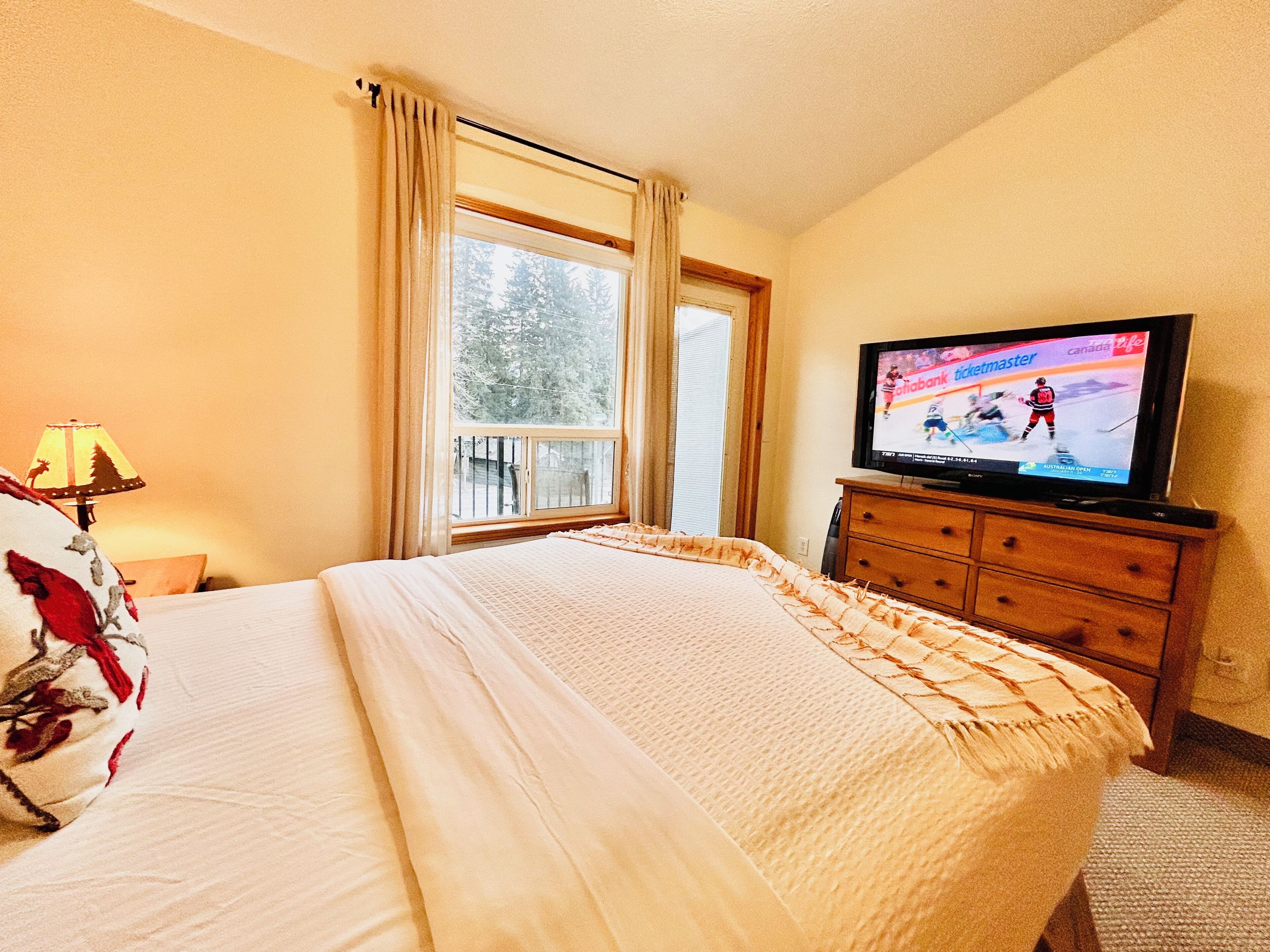 A hotel room with a bed, a television displaying a hockey game, a window with curtains, and a lamp.