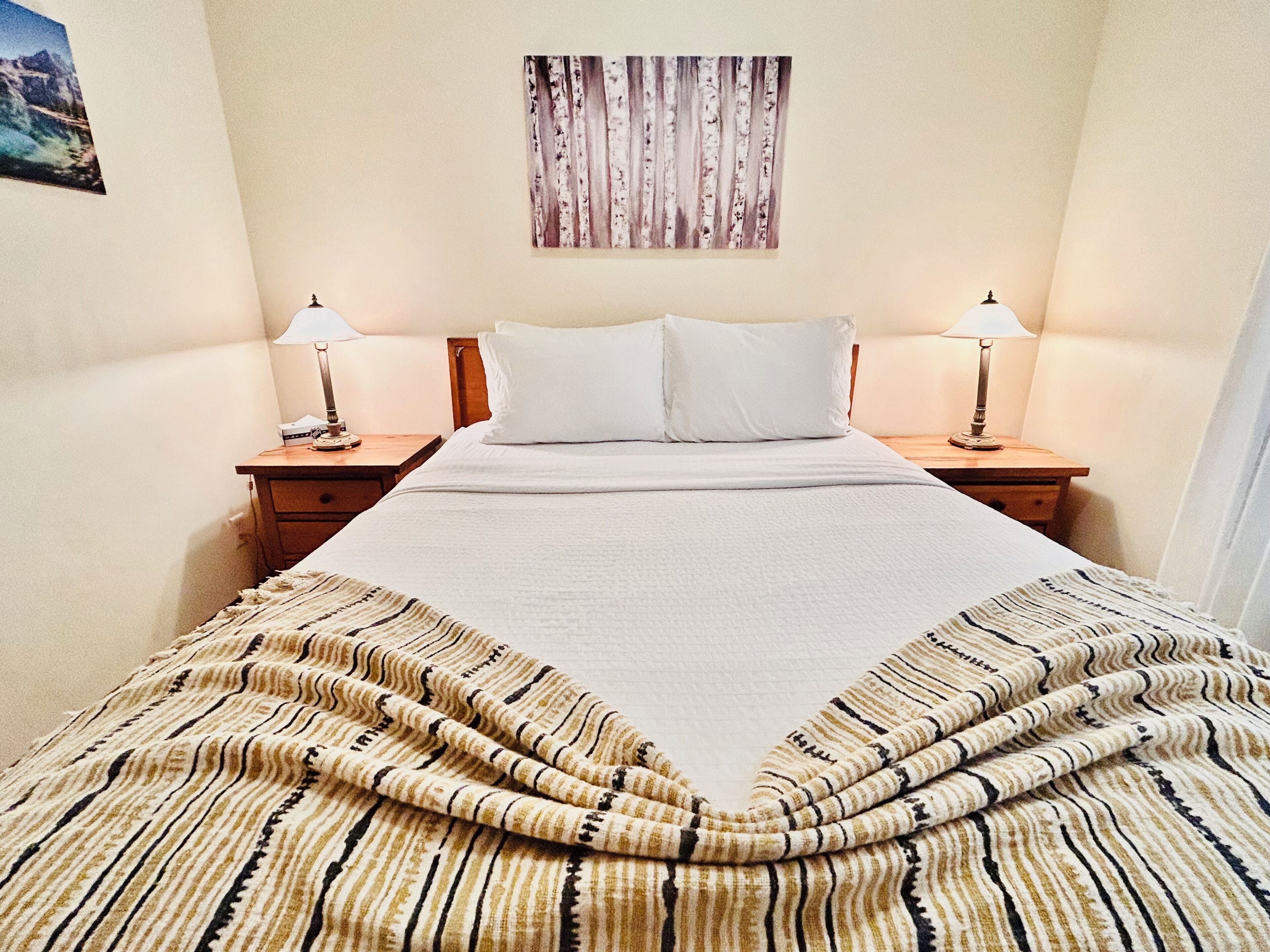 Una cama bien hecha con sábanas blancas, dos mesitas de noche con lámparas, una obra de arte en la pared y un plaid a rayas.