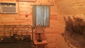 A wooden cabin room with a bed, a nightstand, a lamp, a fur rug, and a wall-mounted heater.