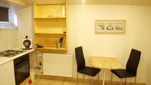 Et kompakt køkken med komfur, ovn og et lille spisebord med to stole. Der er en radiator under hylderne og et billede i ramme på væggen.