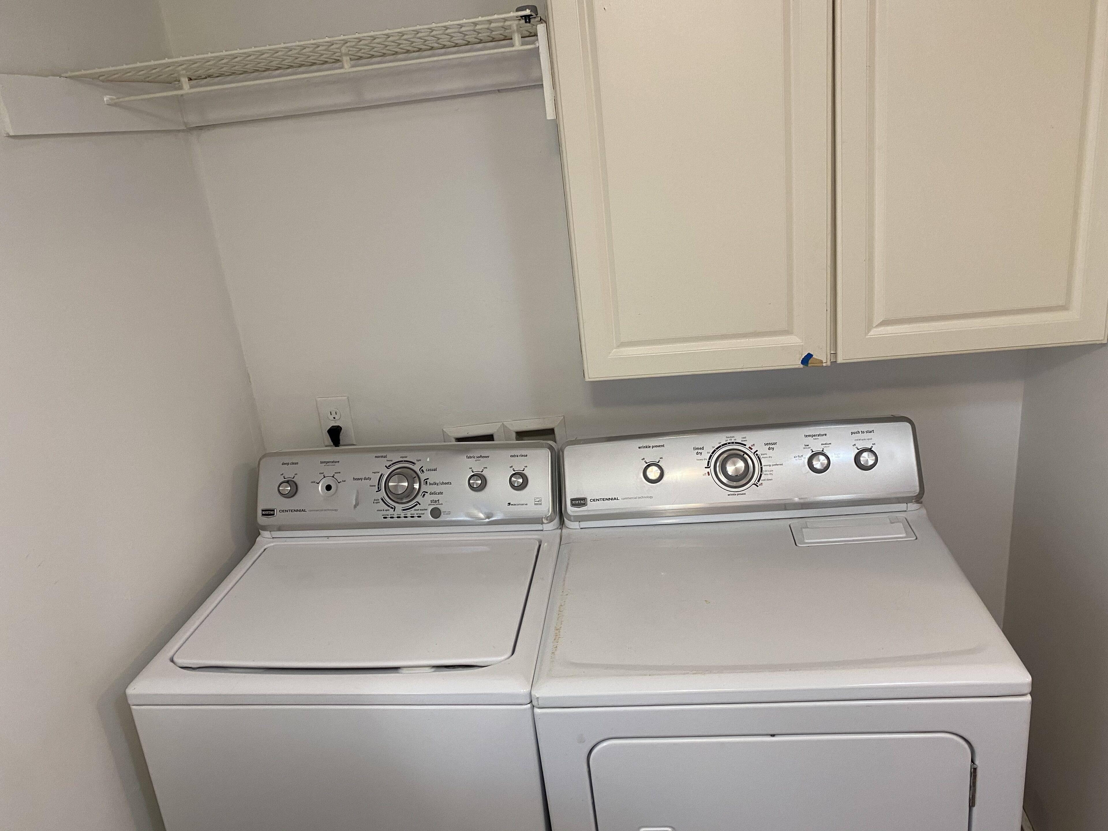 Une buanderie équipée de deux machines à laver et deux sèche-linge superposés, avec un mur blanc et une étagère au-dessus des sèche-linge.