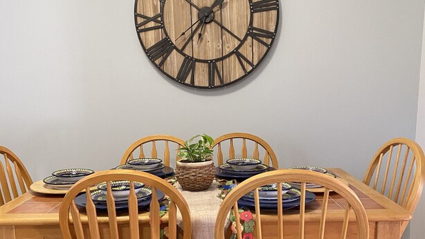 A dining area with a wooden table and chairs, a large wall clock, and a woven basket with a plant.