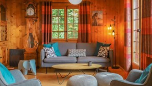 Interior acolhedor de uma cabana com sofá, pufes, mesa de centro redonda, relógio e um quadro na parede.