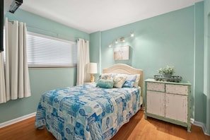 Quarto com cama, mesa de cabeceira, luminária, cômoda e janela com cortinas.