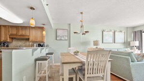 A kitchen with wooden cabinets, a dining table with chairs, a chandelier, and a living area with a sofa and a lamp. - Beautiful ocean & beach views, oceanfront condo w/pool, sundeck, family friendly-North Myrtle Beach (North Myrtle Beach)