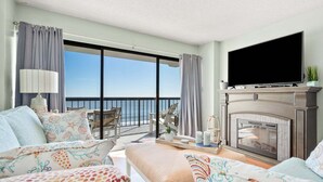 A spacious living room with a large television, a fireplace, and a balcony with a view of the ocean. - Beautiful ocean & beach views, oceanfront condo w/pool, sundeck, family friendly-North Myrtle Beach (North Myrtle Beach)