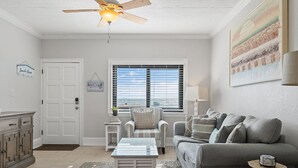 Un salon avec un ventilateur de plafond, un canapé, une table basse et une fenêtre équipée de stores.