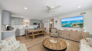 A modern living room with a wooden coffee table, a TV mounted on the wall, and a kitchen area with grey cabinets and a dining table. - Siesta Key Village Heated pool (Siesta Key)