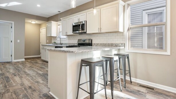 A modern kitchen with white cabinets, a granite countertop, a stainless steel microwave, and a built-in oven. There are two bar stools and a window with blinds.