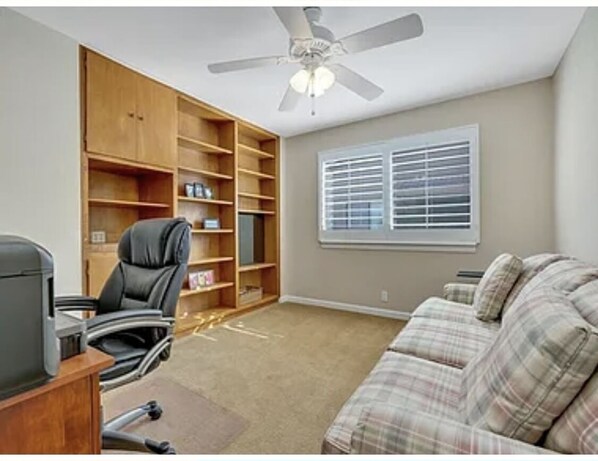 A room with a ceiling fan, a large wooden bookshelf, a black office chair, a plaid sofa, and a window with blinds. - Great resort living in Palm Desert (Palm Desert)
