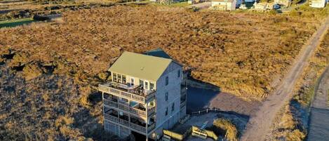 Un edificio de varios pisos con techo verde, situado en una zona costera con camino de tierra y vegetación escasa.