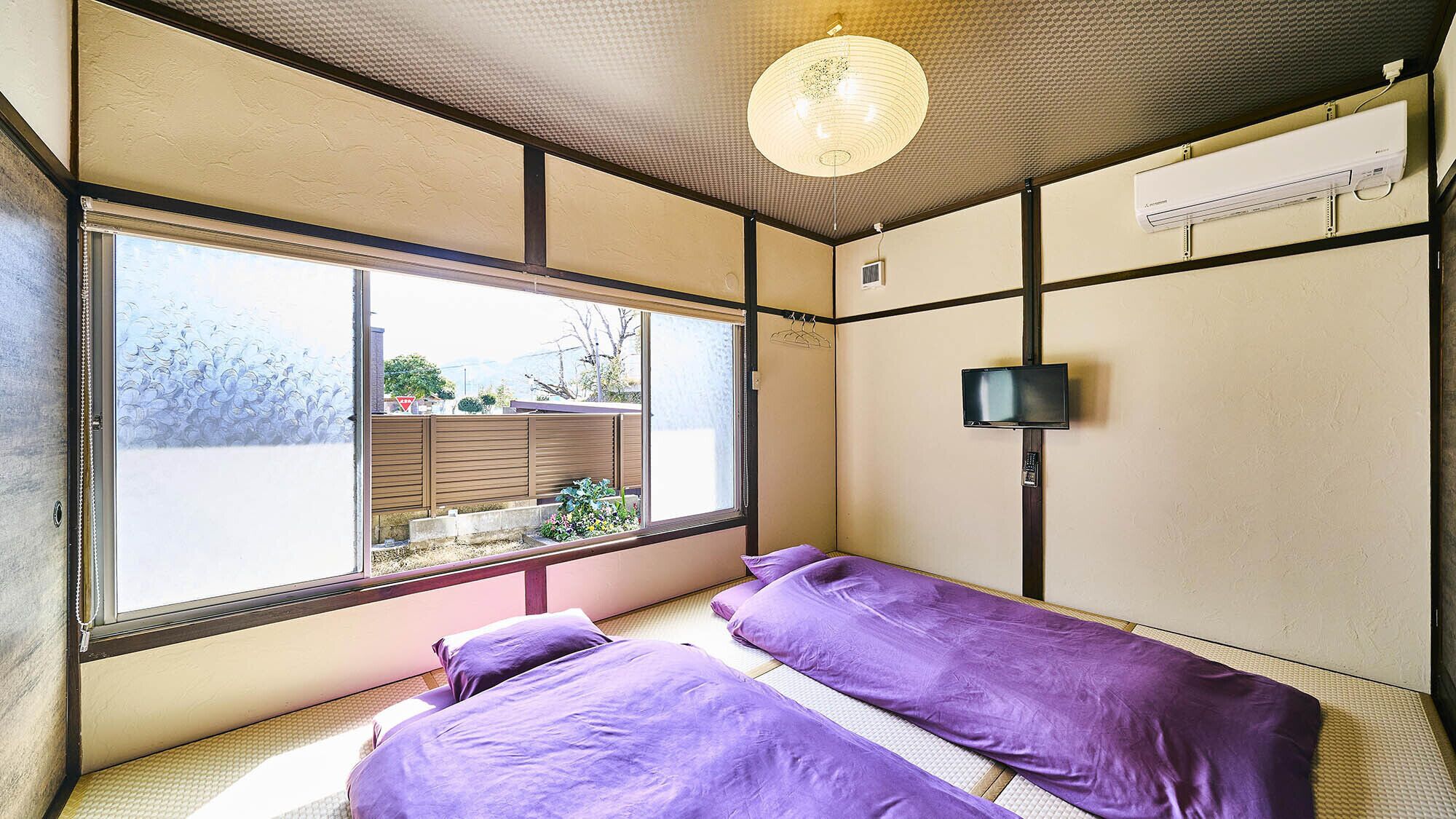 A room with a large window, a bed with purple bedding, a television on the wall, and an air conditioning unit.