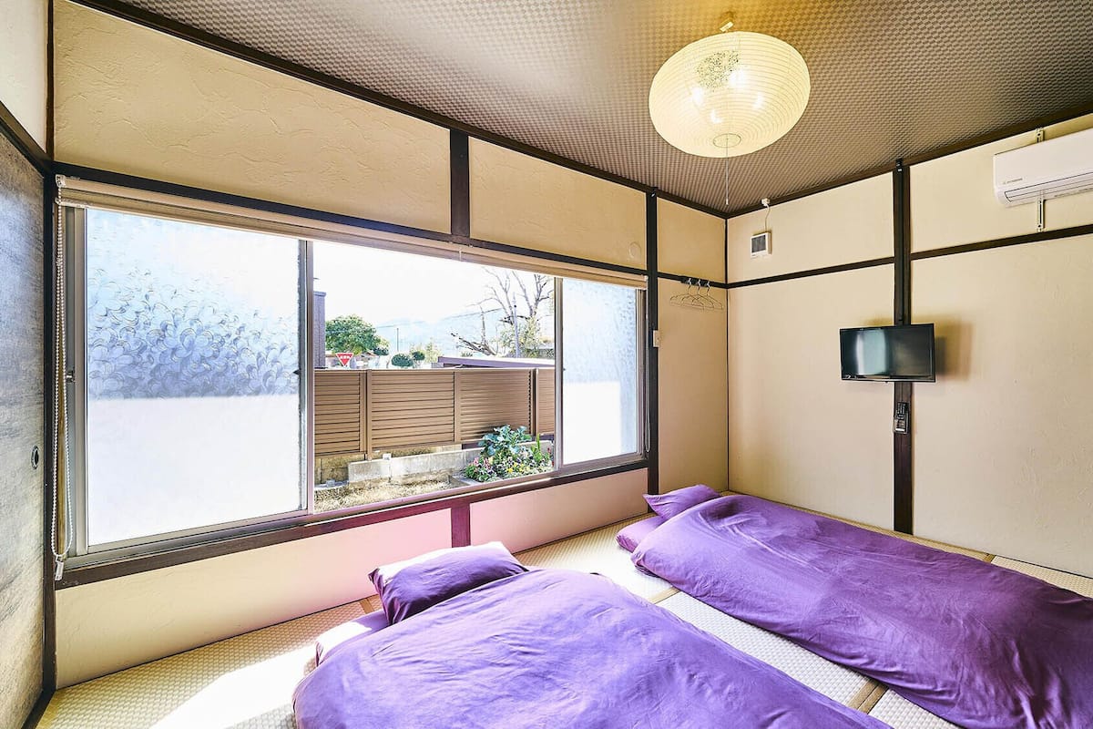 A room with a large window, a bed with purple bedding, a television on the wall, and an air conditioning unit.