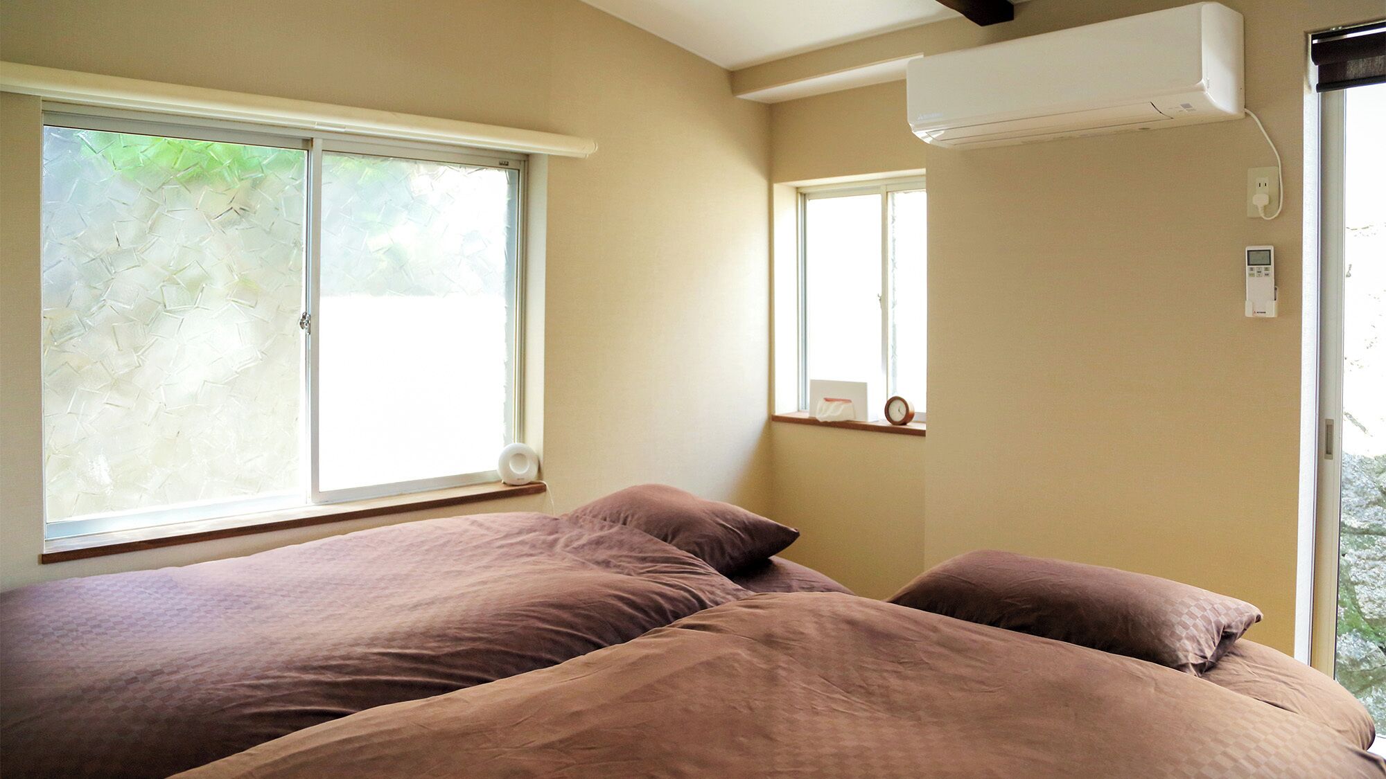 A bedroom with two beds, a window, an air conditioning unit, and a clock on the wall.