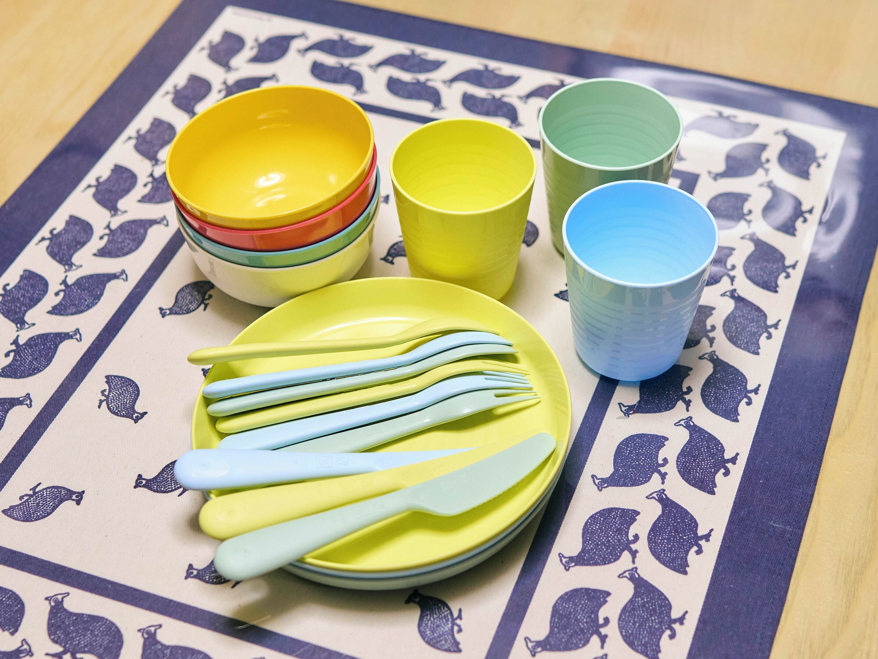 A table with a patterned tablecloth, a stack of colorful bowls, a plate with blue utensils, and a blue cup.