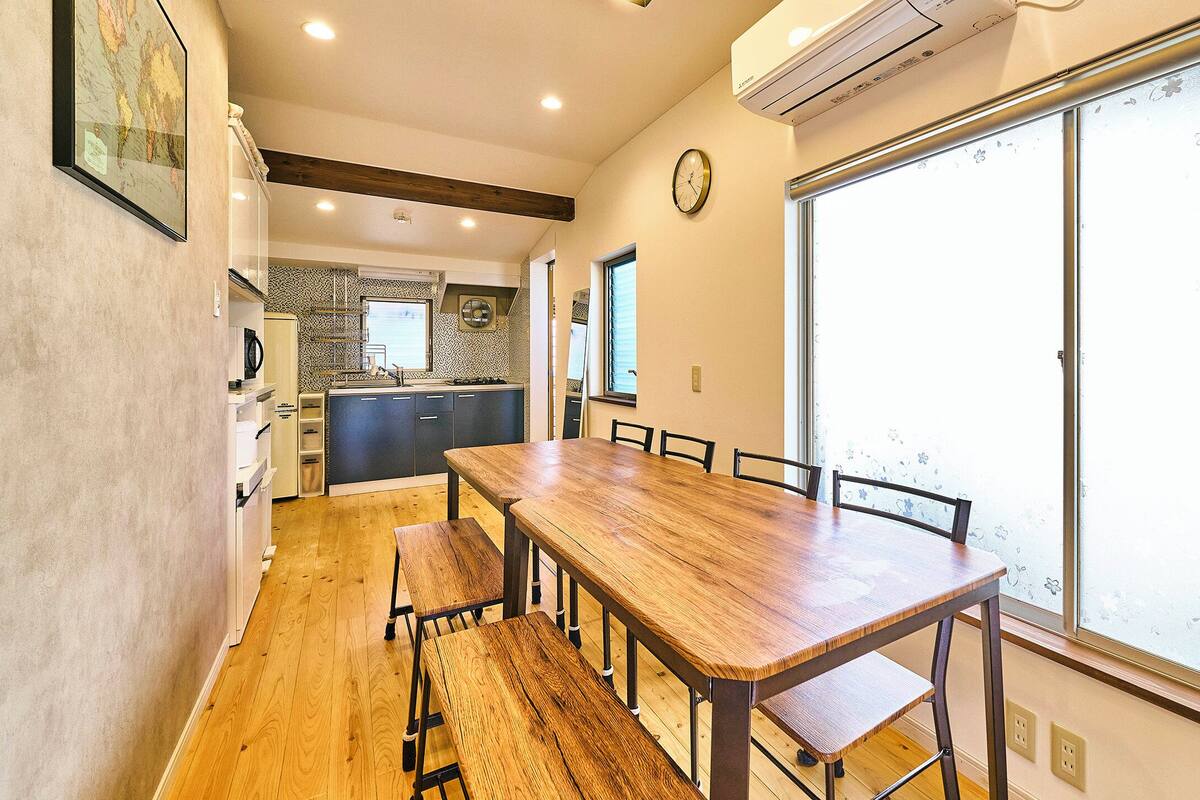 A dining area with a wooden table and benches, a kitchen in the background, and a large window with frosted glass.