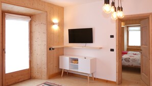 Una habitación con paredes de madera, un televisor de pantalla plana montado en la pared, un mueble blanco para televisor y una puerta que da a otra habitación.